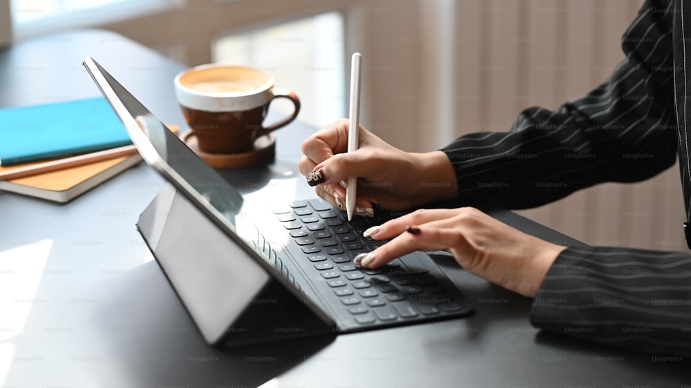 Foto cortada de mulher de negócios digitando no teclado do tablet e segurando a caneta elétrica na mão enquanto se senta na mesa de trabalho moderna com a confortável sala de escritório como fundo.