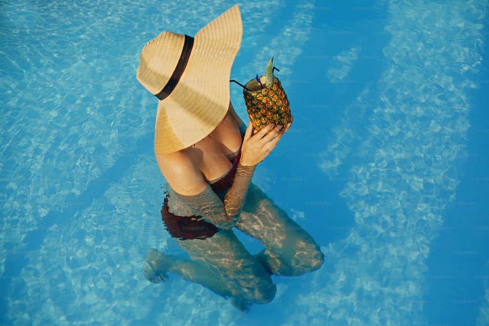 Beautiful young woman in hat holding  cocktail in pineapple and relaxing in pool, summer vacation. Girl enjoying warm sunshine, swimming in pool on rooftop in luxury tropical resort