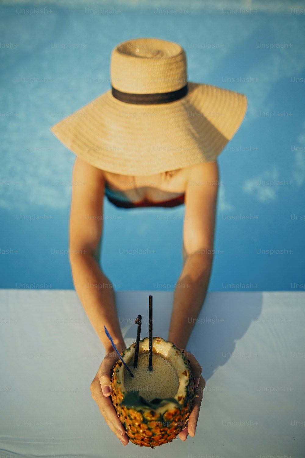 Hermosa joven con sombrero sosteniendo un delicioso cóctel en piña y relajándose en la piscina, vacaciones de verano. Chica disfrutando del cálido sol en la piscina en la azotea en un resort tropical de lujo. Vista superior