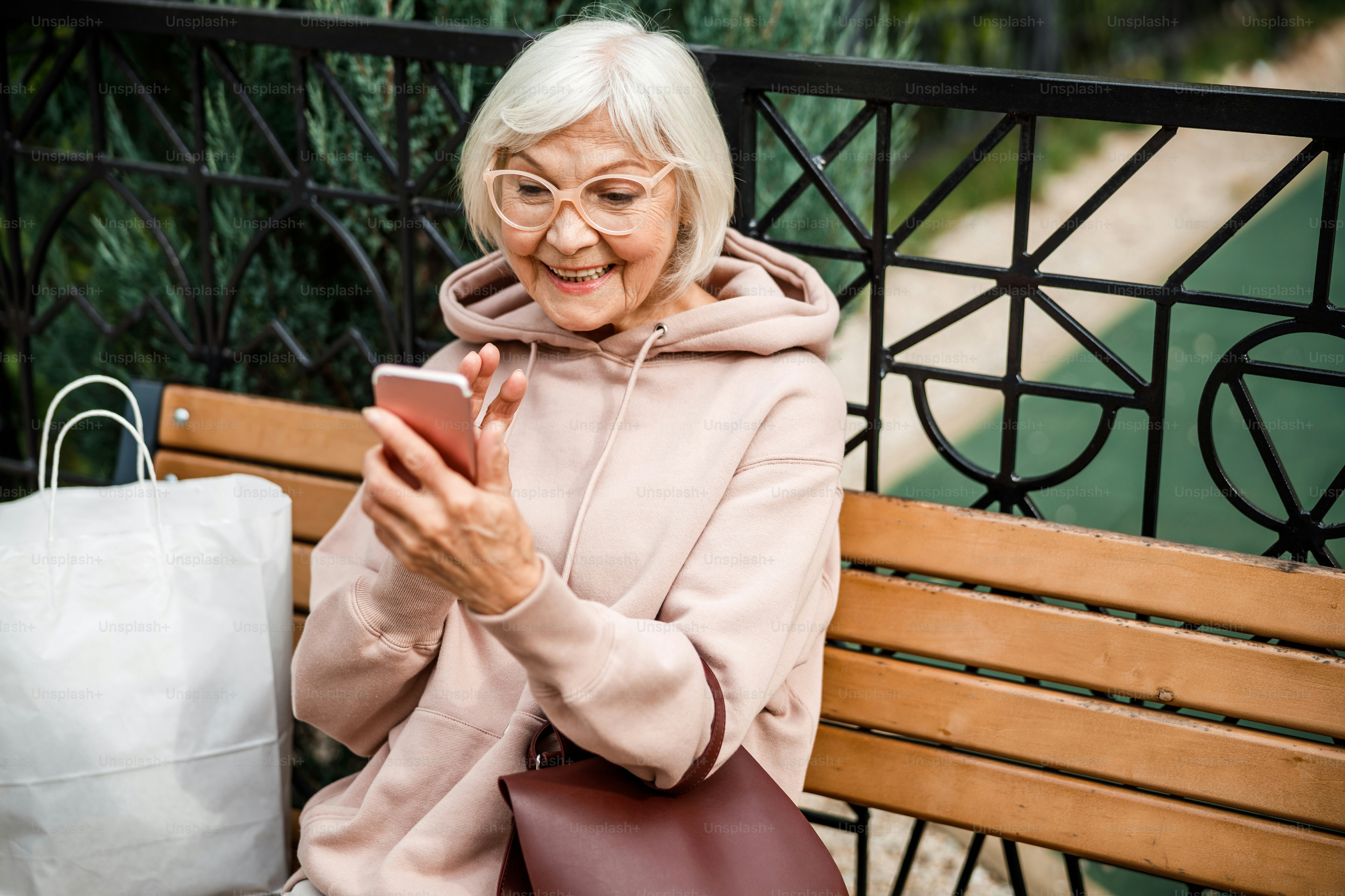 Merry stylish seniour female is sitting on bench in park and ringing using camera on smartphone