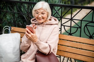 메리 세련된 노인 여성은 공원의 벤치에 앉아 스마트 폰의 카메라를 사용하여 울린다