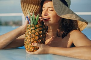 Beautiful young woman enjoying cocktail in pineapple, relaxing in pool on sunny summer vacation. Portrait of girl in hat with natural skin drinking cocktail on rooftop in luxury tropical resort