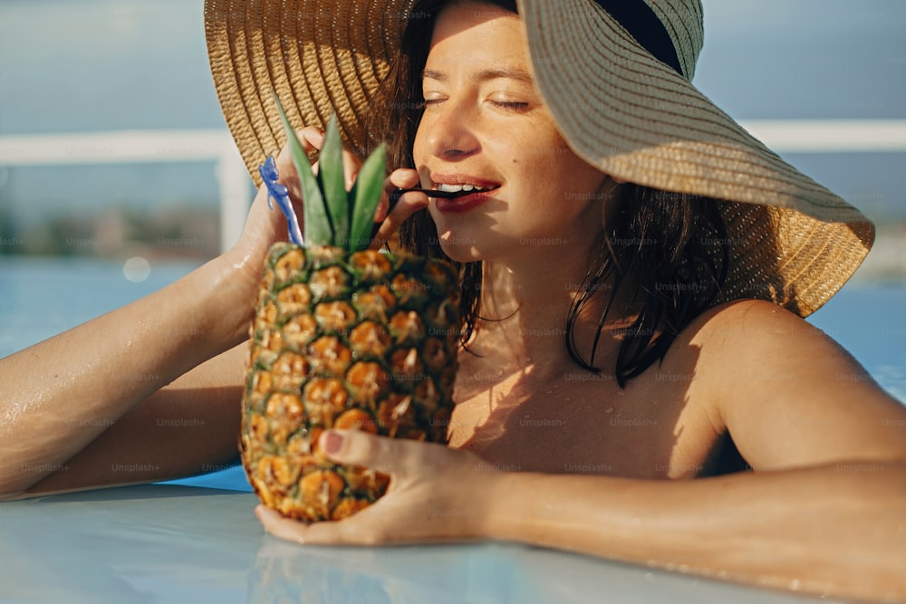 Beautiful young woman enjoying cocktail in pineapple, relaxing in pool on sunny summer vacation. Portrait of girl in hat with natural skin drinking cocktail on rooftop in luxury tropical resort