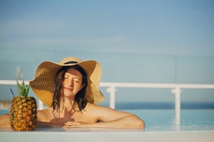 Beautiful young woman in hat relaxing in pool with delicious cocktail in pineapple. Summer vacation. Girl enjoying warm sunshine in pool on rooftop in luxury tropical resort. Copy space