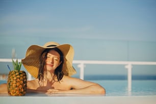 Hermosa joven con sombrero relajándose en la piscina con un delicioso cóctel en piña. Veraneo. Chica disfrutando del cálido sol en la piscina en la azotea en un resort tropical de lujo. Espacio de copia