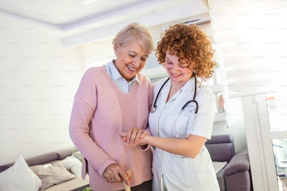 Smiling nurse helping senior lady to walk around the nursing home. Portrait of happy female caregiver and senior woman walking together at home. Professional caregiver taking care of elderly woman.