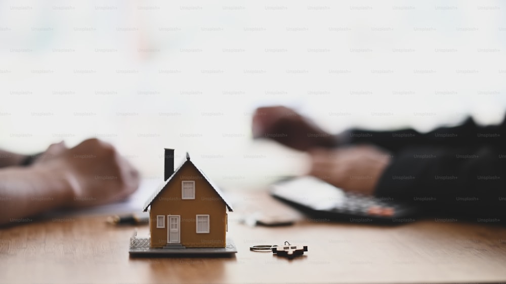 Foto des Hausmodells und des Schlüssels auf dem modernen Holzschreibtisch mit der Hand der Immobilienagentur und des Kunden als Hintergrund.