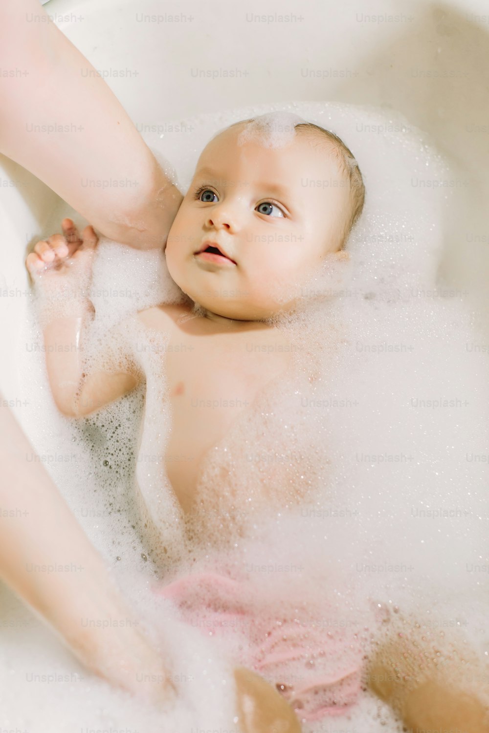 Happy laughing baby taking a bath playing with foam bubbles. Little child in a bathtub. Infant washing and bathing. Hygiene and care for young children. Newborn baby bathing