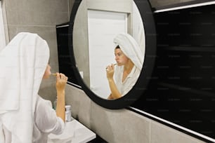 Hermosa mujer joven con toalla blanca cepillándose los dientes con cepillo de dientes de bambú y pasta de dientes de carbón en el baño moderno, mirando el espejo redondo. Higiene dental. Residuo cero.