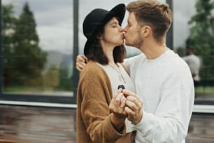 Stylish hipster couple holding together key from their new home on background of terrace and big windows outdoors. Happy young family kissing, celebrating buying first property.