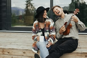 Happy young family travelers smiling and having fun. Hipster man playing on ukulele for his stylish woman, relaxing on wooden porch  of modern cabin with big windows in mountains.