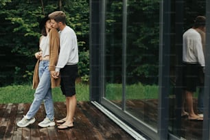 スタイリッシュなヒップスターのカップルが木製のテラスで抱き合ってキスをし、山のキャビンでリラックスしています。大きな窓と木の背景に抱擁するモダンな衣装の幸せな若い家族
