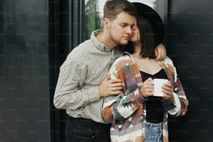 Stylish hipster couple with morning coffee kissing on background of modern cabin with big windows in mountains. Happy young family embracing and enjoying new home in woods. Travel