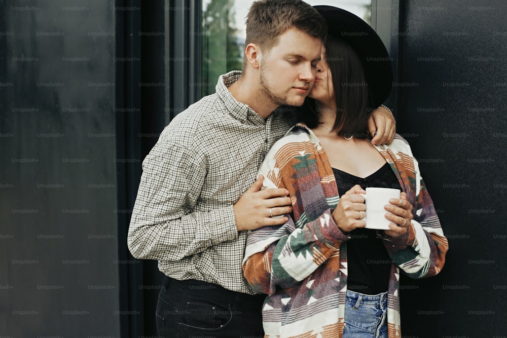 Stylish hipster couple with morning coffee kissing on background of modern cabin with big windows in mountains. Happy young family embracing and enjoying new home in woods. Travel