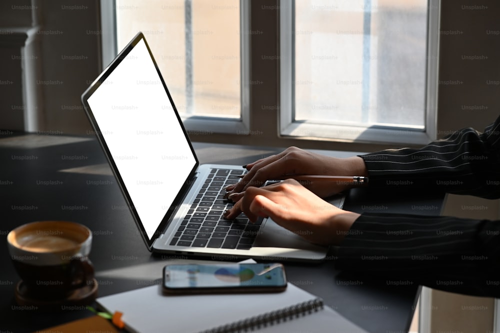 Side shot of young businesswoman typing on white blank screen laptop while sitting at the modern working desk. Coffee cup, smartphone with graphic chart on display putting together on table.