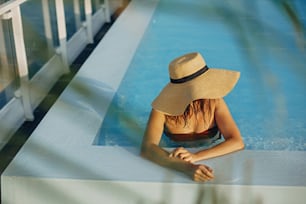 Stylish young woman in hat relaxing in blue pool and enjoying summer holiday. Summer tropical vacation. Girl in sunhat on vacation in luxury resort, swimming in pool on rooftop. Space for text