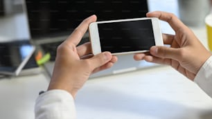 Photo of young businessman's hand holding white smartphone with black blank screen at modern working table with blurred laptop and office equipment as background.
