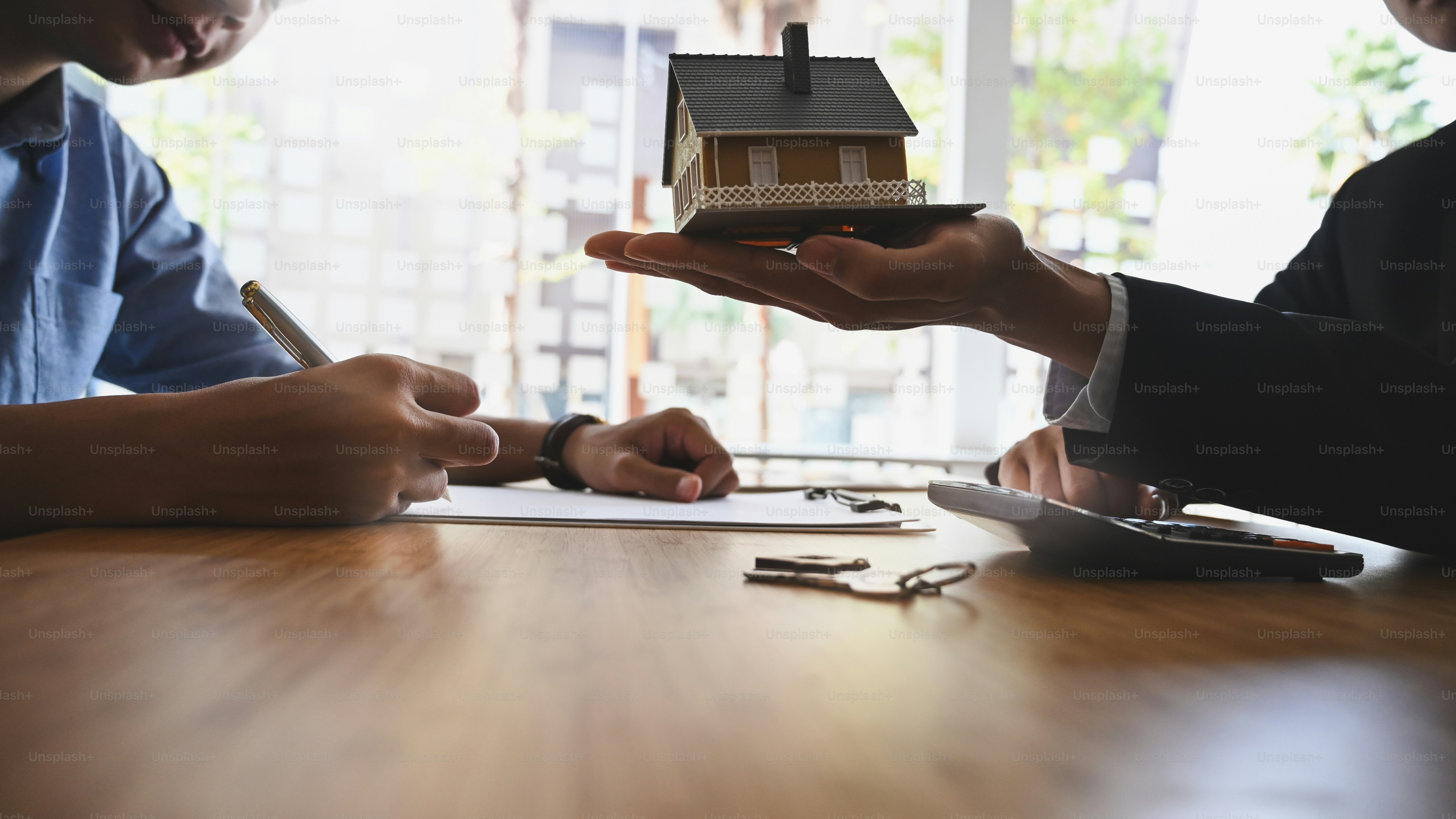 Cropped shot of House broker holding the sample house model in hand with his customer while writing on paper/ signing an agreement at the wooden table. Loan,Debt, selling/buying agreement concept.