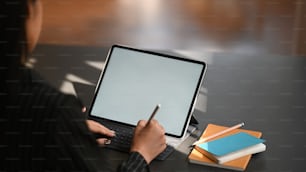 Cropped shot through shoulder of business woman typing on keyboard of tablet and holding electric pen in hand while sitting at the modern working table with the comfortable office room as background.