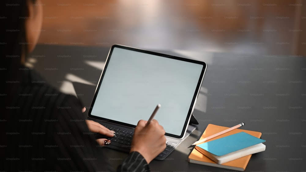 Cropped shot through shoulder of business woman typing on keyboard of tablet and holding electric pen in hand while sitting at the modern working table with the comfortable office room as background.