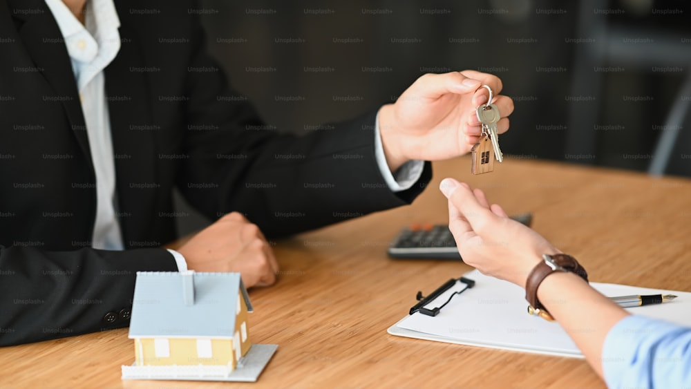 Cropped shot of House broker giving a property key to his customer while he has writing/signing on agreement at the modern wooden desk. Loan, Debt, Credit, Buying or Selling agreement concept.