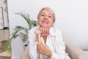 白い髪と杖を持つ美しい年配の女性の肖像画。自宅のソファに座っている年配の女性の肖像画。カメラを見ている笑顔の中年の成熟した白髪の女性