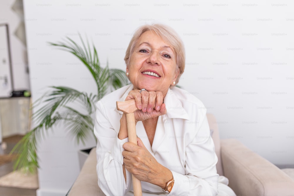 Portrait of beautiful senior woman with white hair and walking stick. Portrait of senior woman sitting on sofa at home. Smiling middle aged mature grey haired woman looking at camera