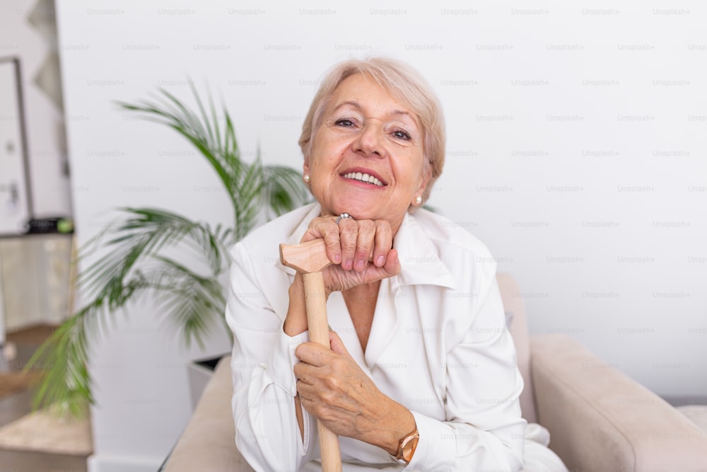 白い髪と杖を持つ美しい年配の女性の肖像画。自宅のソファに座っている年配の女性の肖像画。カメラを見ている笑顔の中年の成熟した白髪の女性