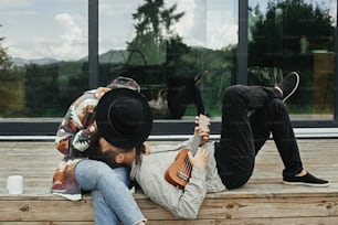 Hipster man playing on ukulele for his beautiful stylish woman, relaxing on wooden porch on background of modern cabin with big windows in mountains. Happy young family travelers enjoying vacation