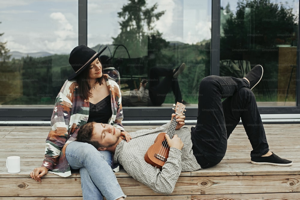 Stylish hipster couple sitting on wooden porch, relaxing with ukulele  on background of modern cabin with big windows in mountains. Happy young family travelers enjoying vacation in woods