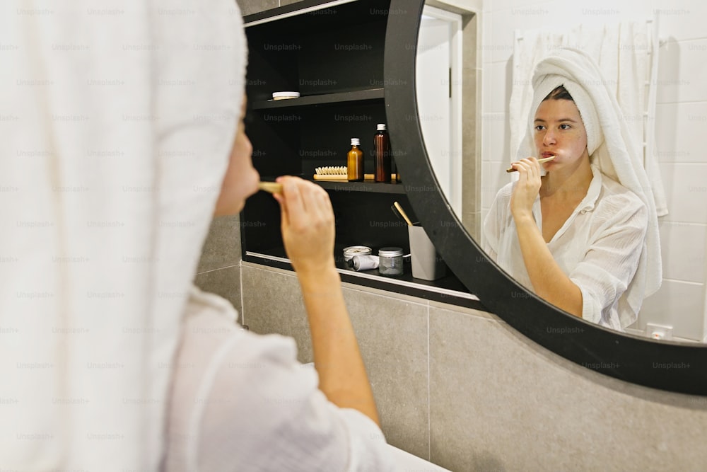 Dental hygiene. Zero waste. Beautiful young woman in white towel brushing her teeth with bamboo toothbrush and charcoal toothpaste in modern bathroom, looking at round mirror.