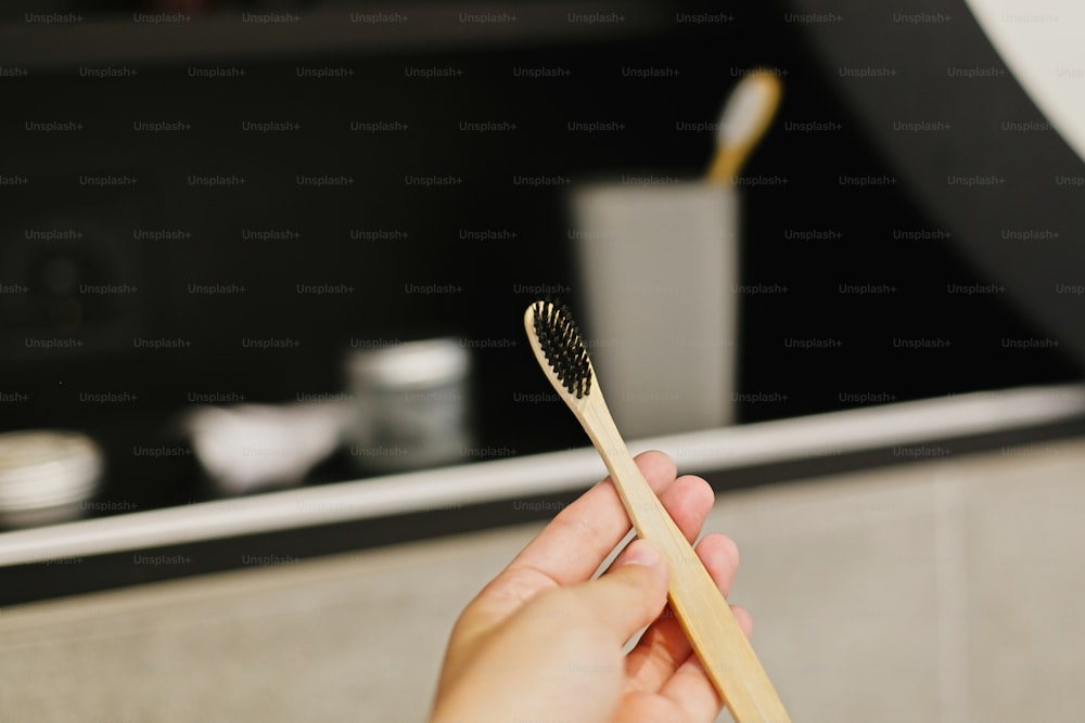 Hand holding natural bamboo toothbrush in modern bathroom on background of stylish black shelf with plastic free natural organic essentials. Zero waste concept. Natural essentials