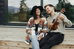 Hipster man playing on ukulele for his stylish woman, relaxing on wooden porch of modern cabin with big windows in mountains. Happy young family travelers smiling and enjoying vacation