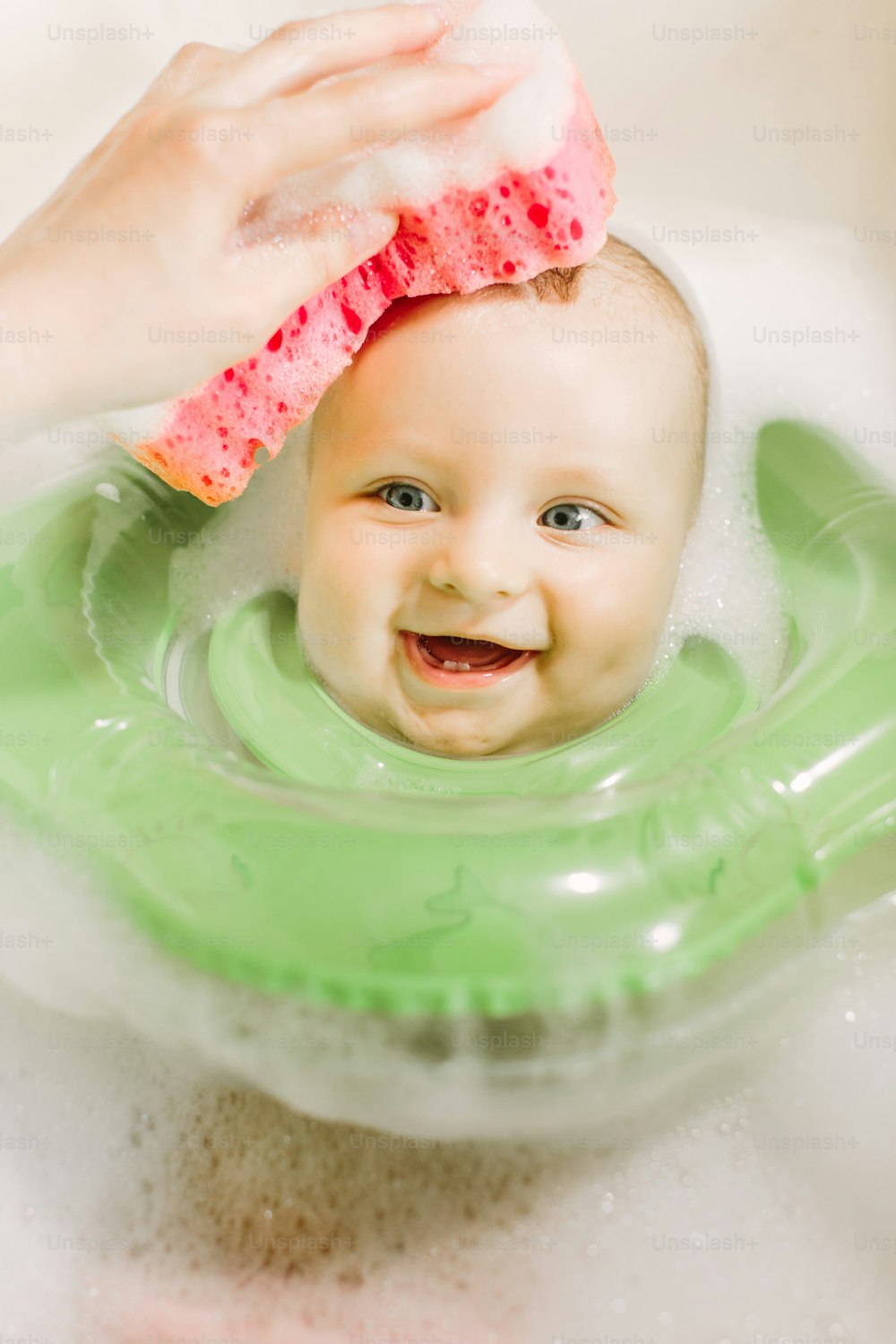 Bebê nadando com anel de natação de pescoço verde. Mãe lavando a cabeça do bebê com uma esponja vermelha