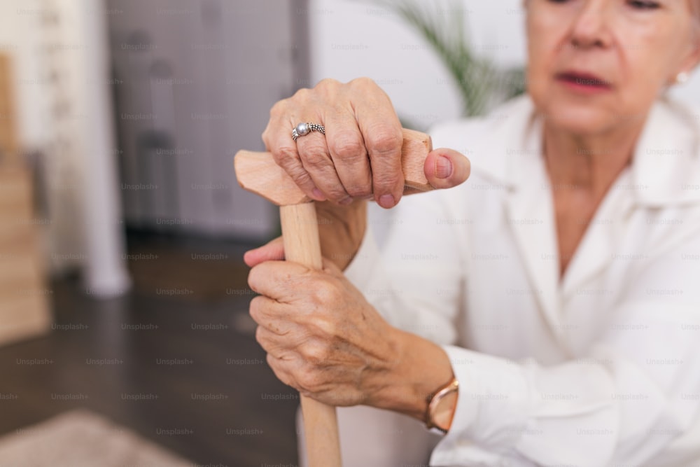 Hände einer alten Frau mit einem Stock, ältere Dame sitzt auf der Couch mit hölzernem Gehstock. Ausschnittaufnahme einer älteren Frau mit einem Stock in einem Altersheim