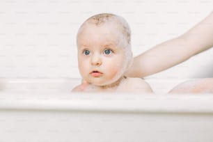 Happy laughing baby taking a bath playing with foam bubbles. Little child in a bathtub. Infant washing and bathing. Hygiene and care for young children.