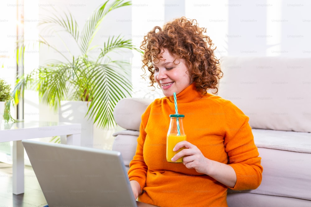 Young smiling red hair woman working from home, she is connecting with a laptop and drinking an healthy orange juice. Cute ginger girl surfing the web on her laptop