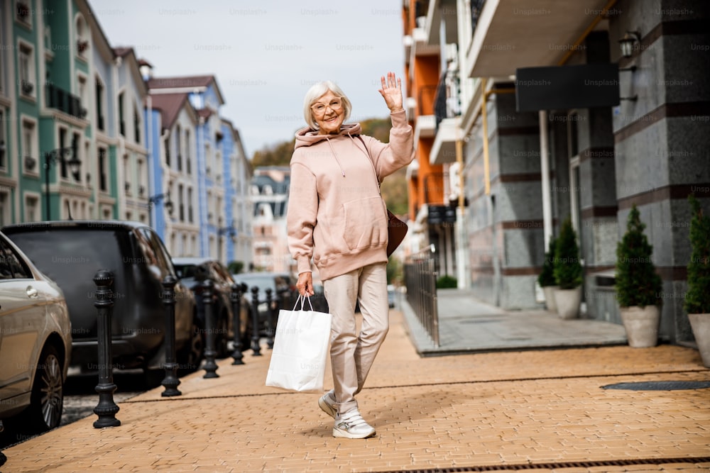 Senhora idosa alegre em roupas esportivas está acenando olá durante a caminhada na rua