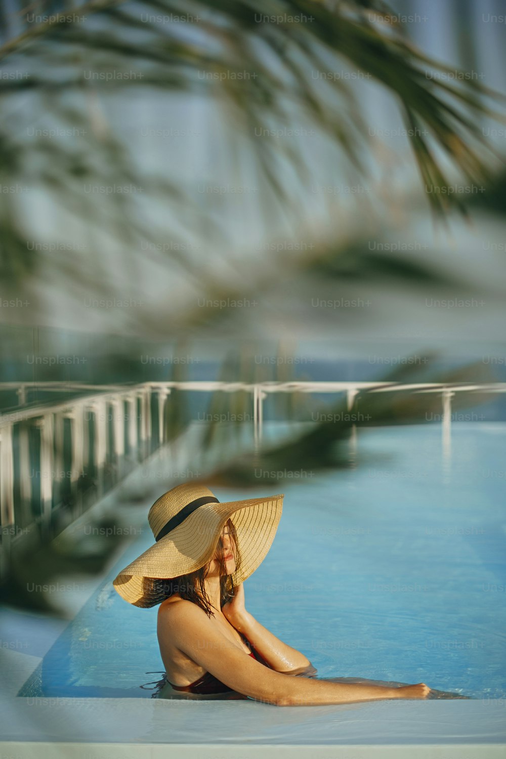 Stylish young woman in hat relaxing in pool under palm leaves and enjoying warm sunny light on summer vacation. Girl in sunhat on vacation in luxury tropical resort, swimming in pool on rooftop