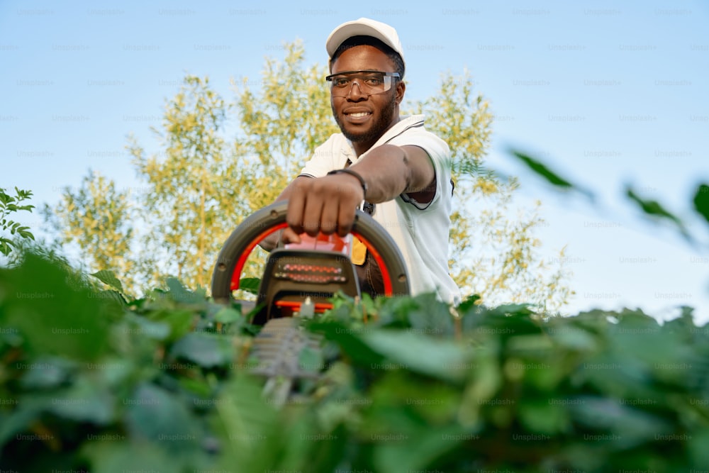 保護メガネと夏の帽子をかぶった陽気なアフリカ人男性の肖像画は、ハンドトリマーで生け垣を剪定しています。晴れた日に茂みを刈る作業服を着た男性の庭師。