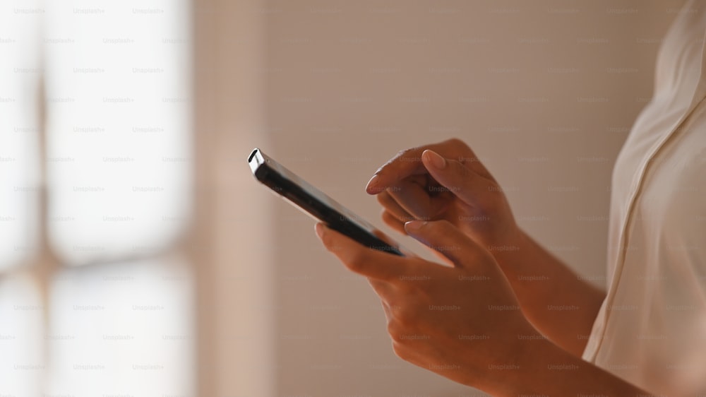 Foto lateral de bela empresária de camisa branca navegando na internet usando smartphone na mão com sala de estar minimalista moderna como fundo.