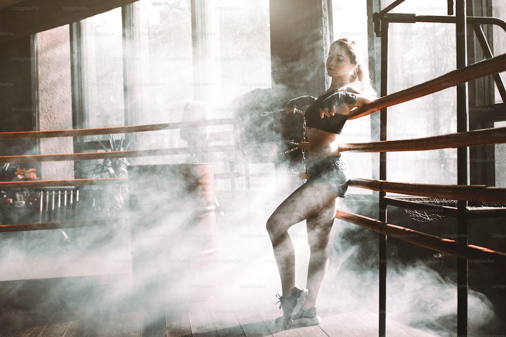 Young pretty boxer woman standing on ring. Full body portrait of boxer woman wearing black sports bra, grey trousers, trainers standing in ring and leaning on ropes. Boxing, sport concept