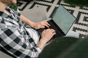 Cropped view of busy young adult guy working at modern laptop computer with copy space on display, using keyboard and sitting in comfort armchair