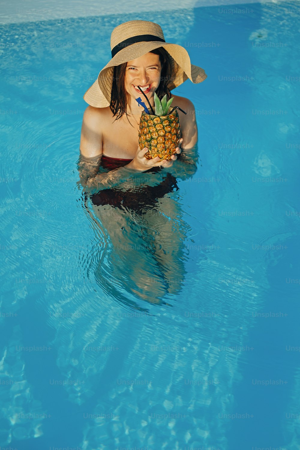 Hermosa joven con sombrero bebiendo un delicioso cóctel de piña y relajándose en la piscina, vacaciones de verano. Muchacha disfrutando de un cálido sol, nadando en la piscina en la azotea en un resort tropical de lujo