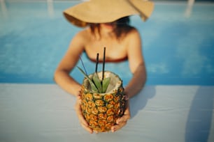 Veraneo. Hermosa joven con sombrero sosteniendo un delicioso cóctel en piña y relajándose en la piscina. Chica disfrutando del cálido sol en la piscina en la azotea en un resort tropical de lujo. Vista superior