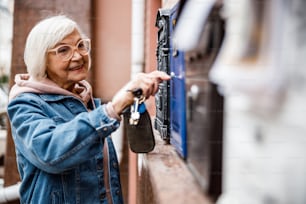 Lächelnde Seniour Frau benutzt Schlüssel zum Öffnen des Briefkastens auf der Straße. Website-Banner