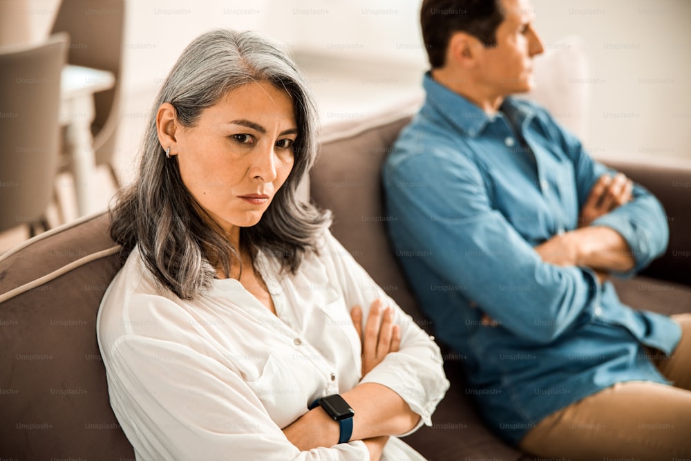 Mature couple having a quarrel while sitting on sofa at home with folded arms looking mad