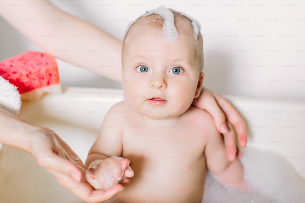 Bambino felice che ride facendo il bagno giocando con le bolle di schiuma. Bambino piccolo in una vasca da bagno. Lavaggio e bagnetto dei neonati. Madre irriconoscibile che fa il bagno al suo bambino in un piccolo pipistrello di plastica bianco