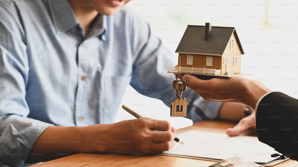Cropped shot of House broker holding the sample house model in hand with his customer while writing on paper/ signing an agreement at the wooden table. Loan,Debt, selling/buying agreement concept.