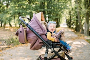 Cute little beautiful baby girl sitting in the pram or stroller and waiting for mom. Happy smiling child with blue eyes